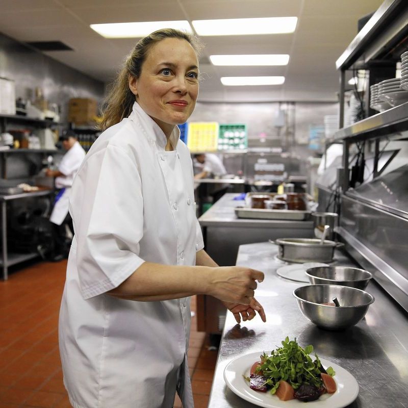 Sarah Stegner wearing a white chef coat in a commercial kitchen plating beets and microgreens on a stainless steel prep table.