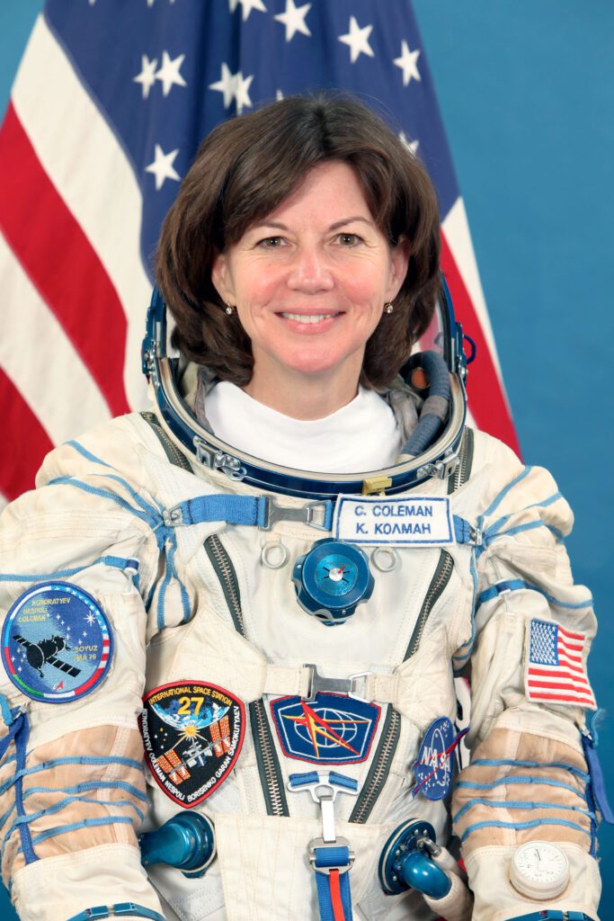 Cady Coleman, smiling, wearing her space suit in front of the U.S. flag.